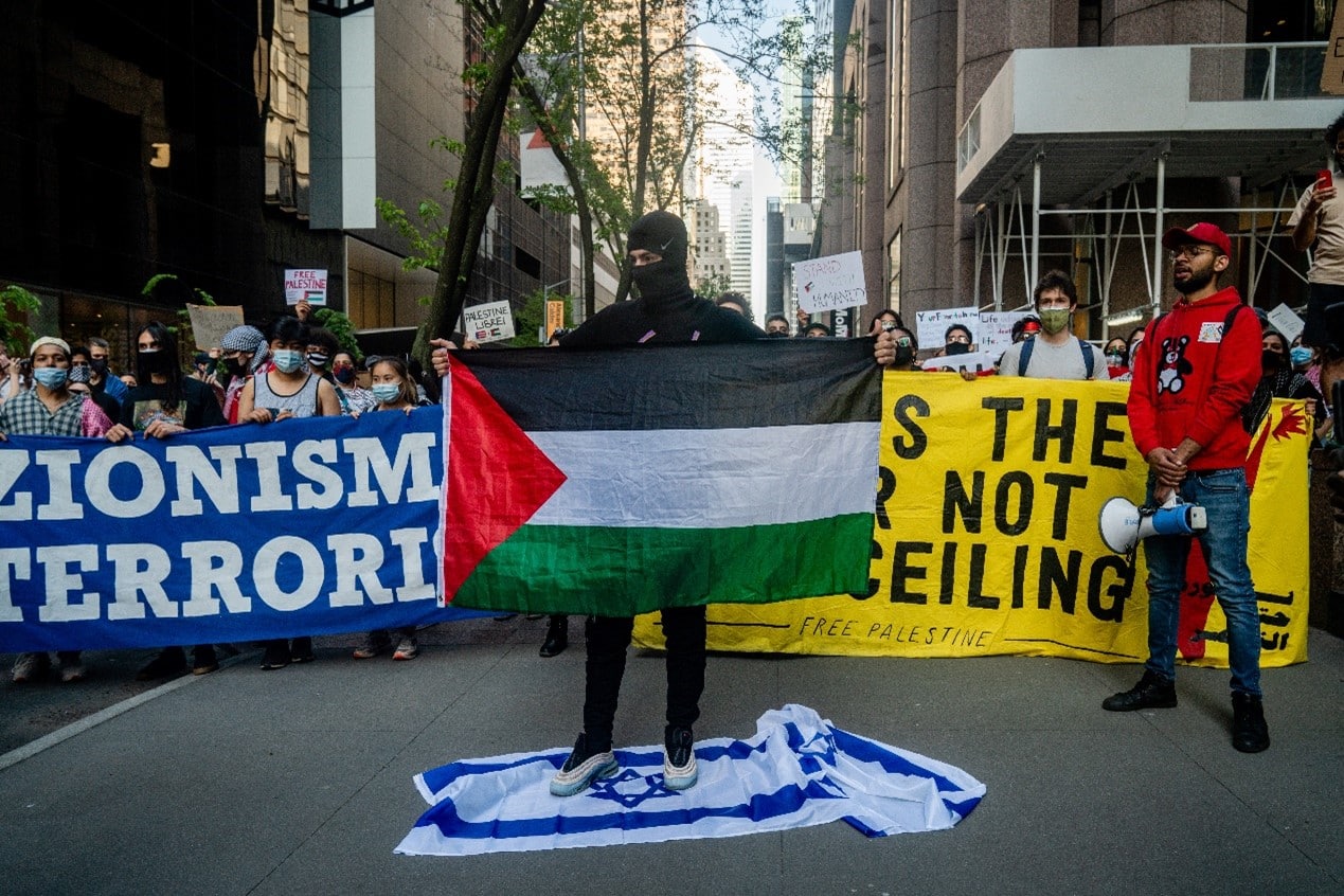 Masked demonstrator holding palestinian flag while standing on Israel flag