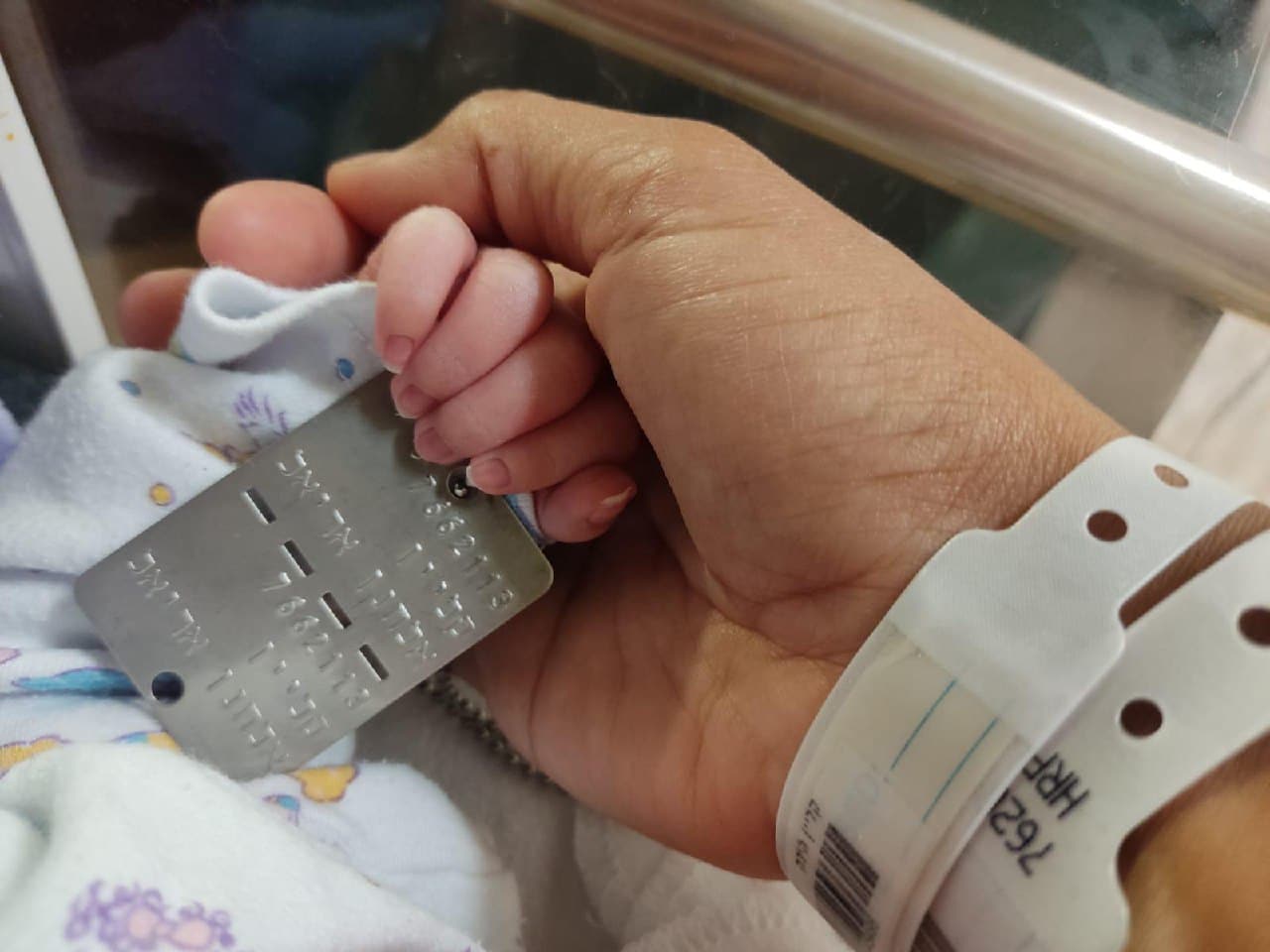 Photo of a hand with a hospital bracelet holding the hand of a newborn baby holding a Klein Elchanan Ariel soldier disk