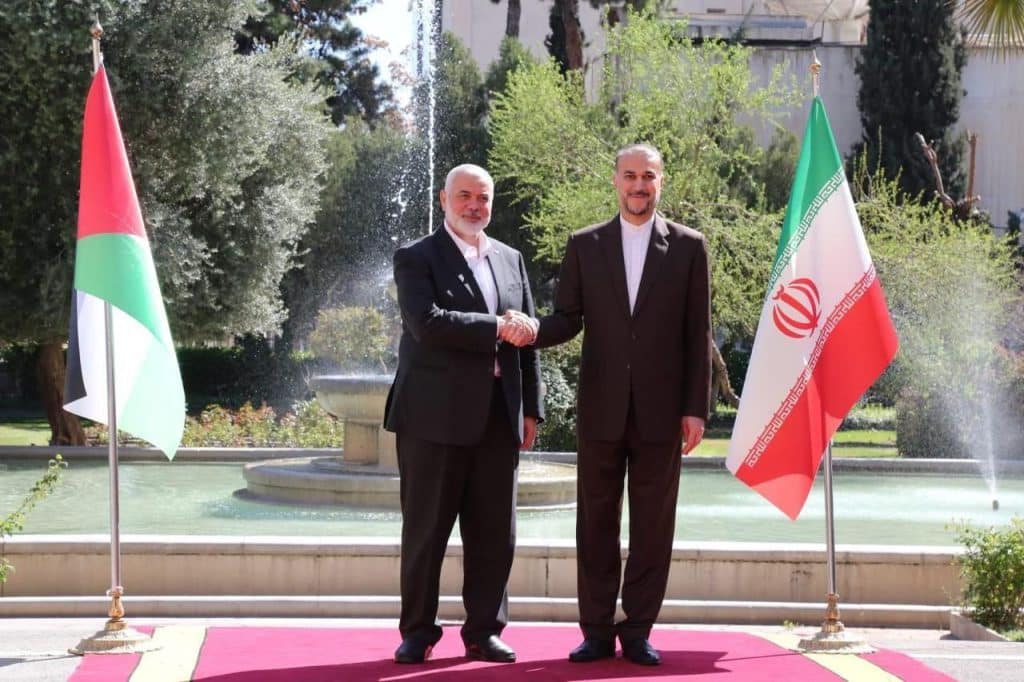 haniyyeh shaking hands with Iranian foreign minister on red carpet between two iranian flags, and a fountain behind