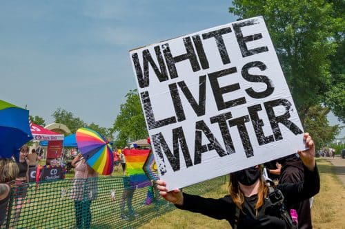 מפגינה רעולת פנים מחזיקה שלט - white lives matter מול יריד עם דגלי גאווה ושמשיות צבעוניות