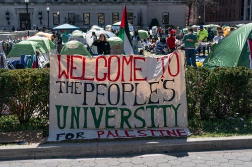 שלט 'ברוכים הבאים לאוניברסיטת הציבור למען פלסטין' וברקע הפגנה בקמפוס עם דגלים פלסטינים