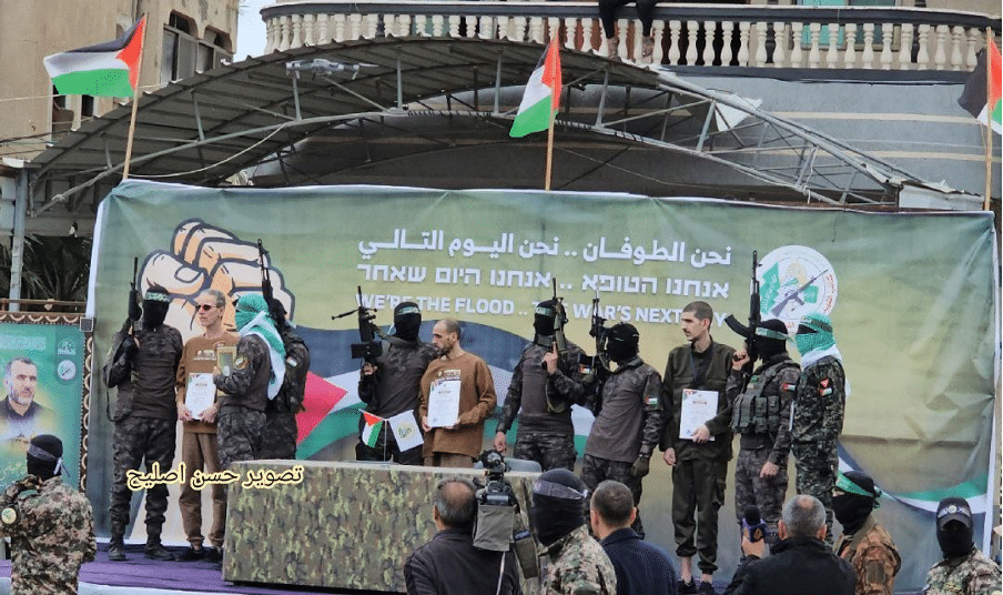 Released hostages on stage with Hamas terrorists given “release” certificates. Hamas wrote on the poster with mistakes in Hebrew and English “We are the flood…The war’s next day” as a message that Hamas would continue fighting. | Source: Hebron News Network on Telegram, https://t.me/khalelnews