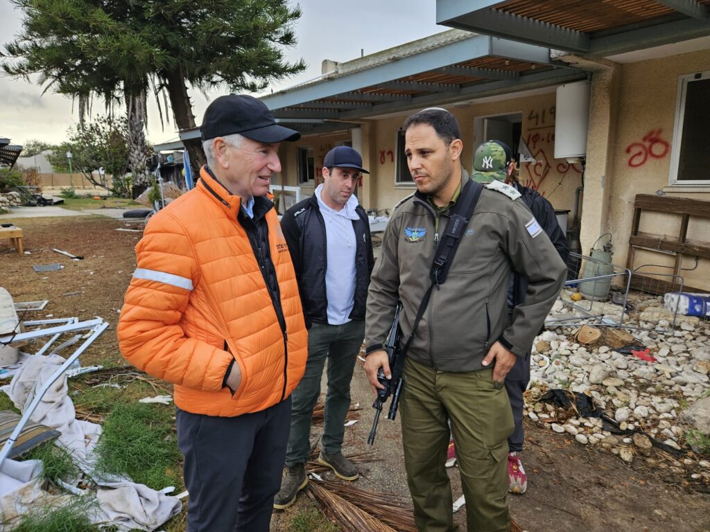 שלושה גברים עומדים ומשוחחים באזור שניזוק, עם מבנים הרוסים וציוד מפוזר ברקע. אחד מהם לובש מעיל כתום, אחד לובש מדים צבאיים ואוחז בנשק, ואדם נוסף עומד ביניהם בלבוש אזרחי. סביבם עדויות ברורות לנזק בזירה.