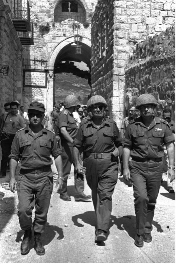צילום שחור-לבן של קציני צה״ל במדי שדה וקסדות, צועדים בסמטה אבן בעיר העתיקה לאחר הקרבות, מוקפים בחיילים ואזרחים.
